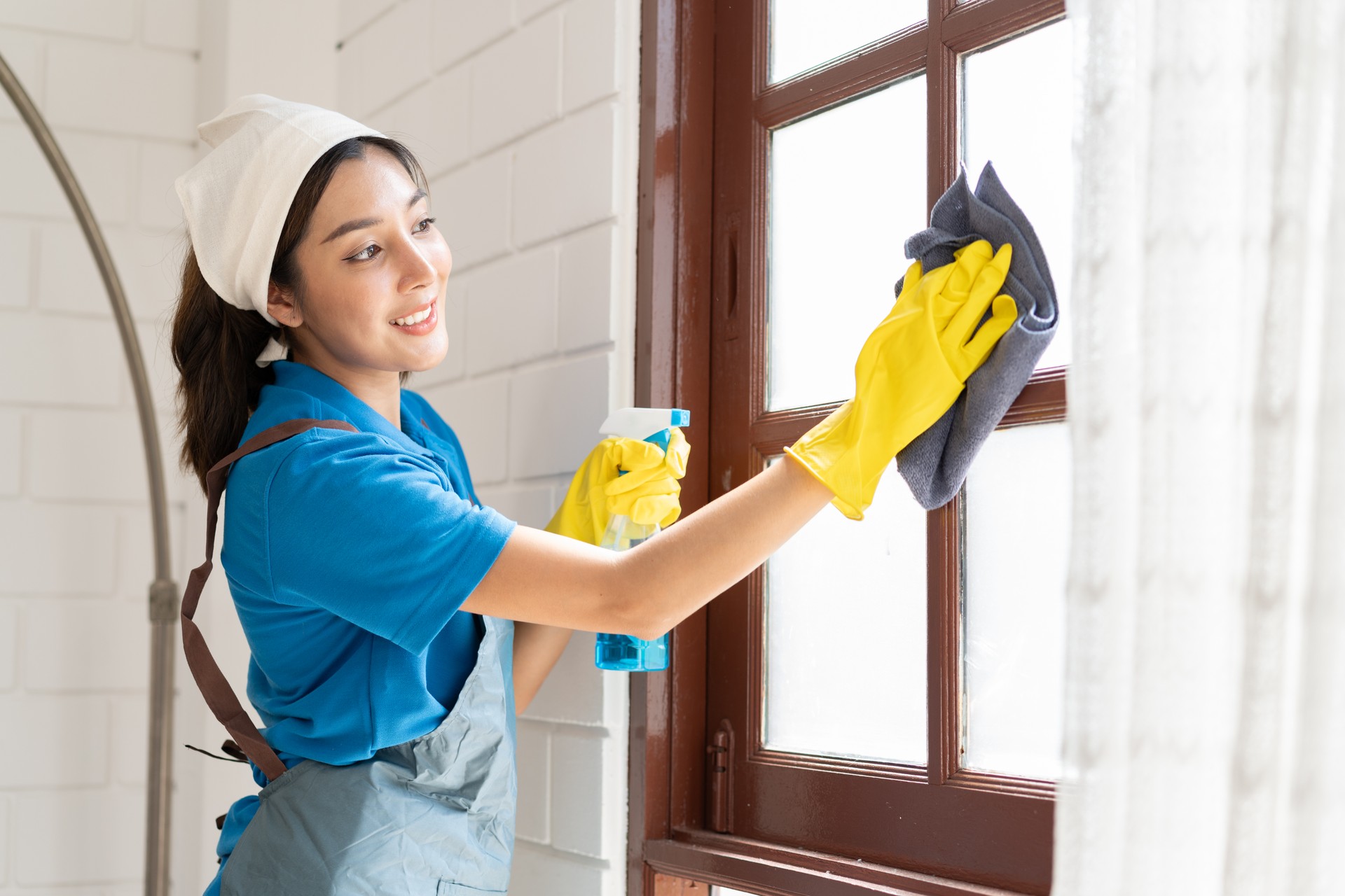 Asia woman in workwear maid holding bottle spray and wiping with microfiber cloth to window at home